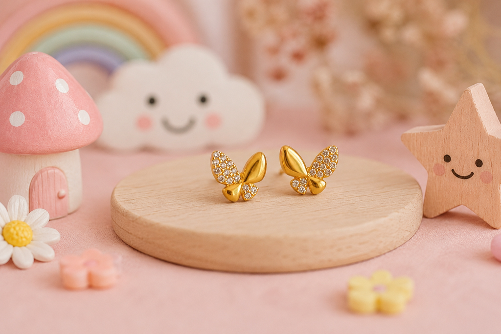 A pair of gold fairy earrings with sparkling crystal wings, displayed on a round wooden stand against a soft pink background with cute decorative props like a smiling cloud, a wooden star, and a small mushroom house.
