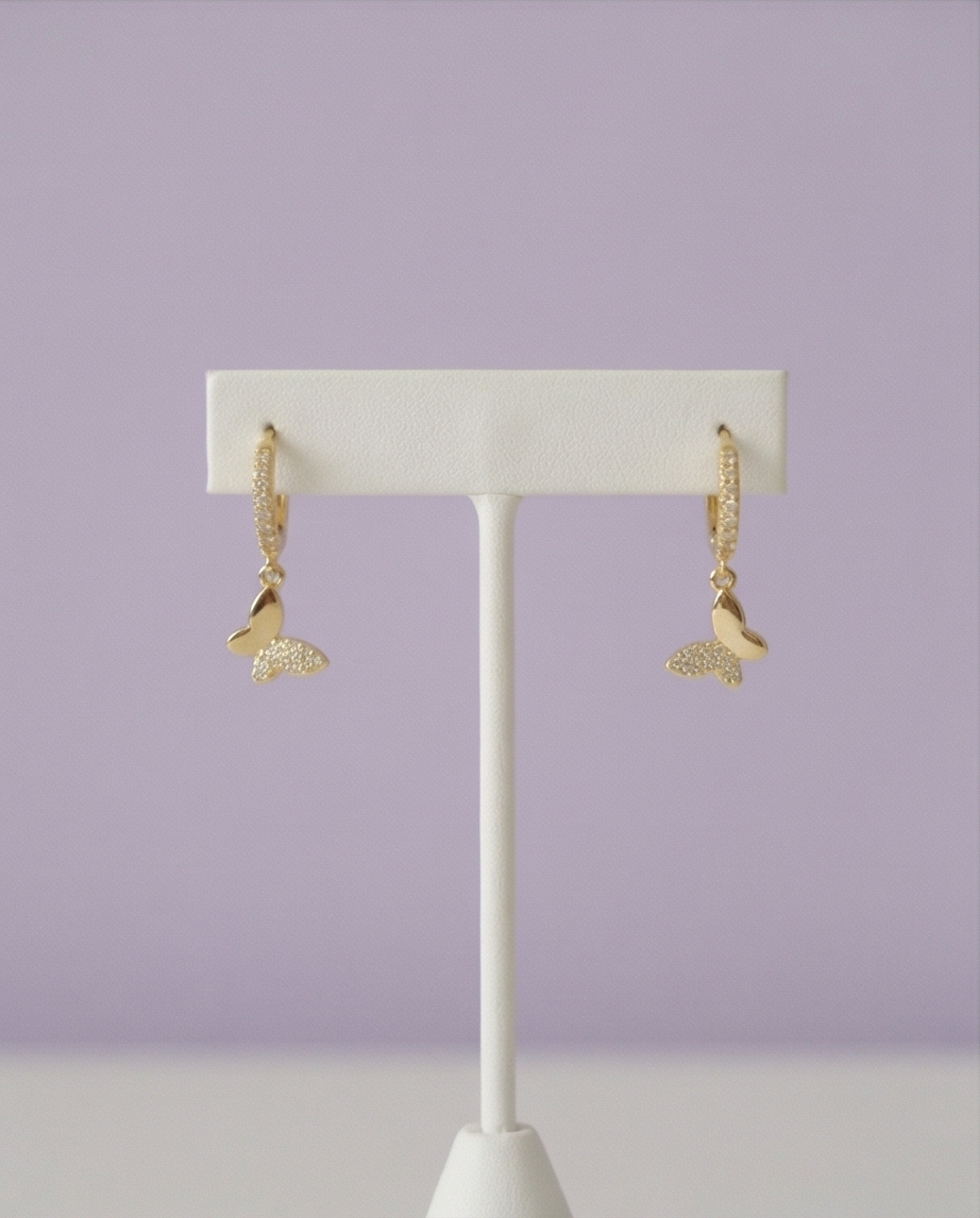A pair of gold dangling earrings with a textured mermaid tail design, displayed on a white T-stand against a soft lavender background.