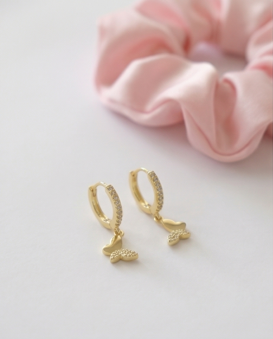 A pair of gold huggie hoop earrings with small dangling textured charms, displayed on a light gray surface with a soft pink fabric scrunchie in the background.