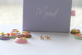 A pair of gold butterfly stud earrings with colorful enamel details, displayed on a white surface in front of a lavender box with the word 'Mood' written on it.