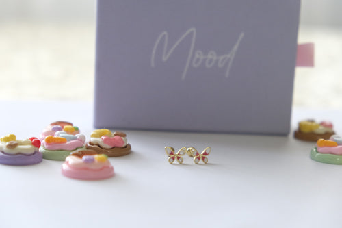 A pair of gold butterfly stud earrings with colorful enamel details, displayed on a white surface in front of a lavender box with the word 'Mood' written on it.