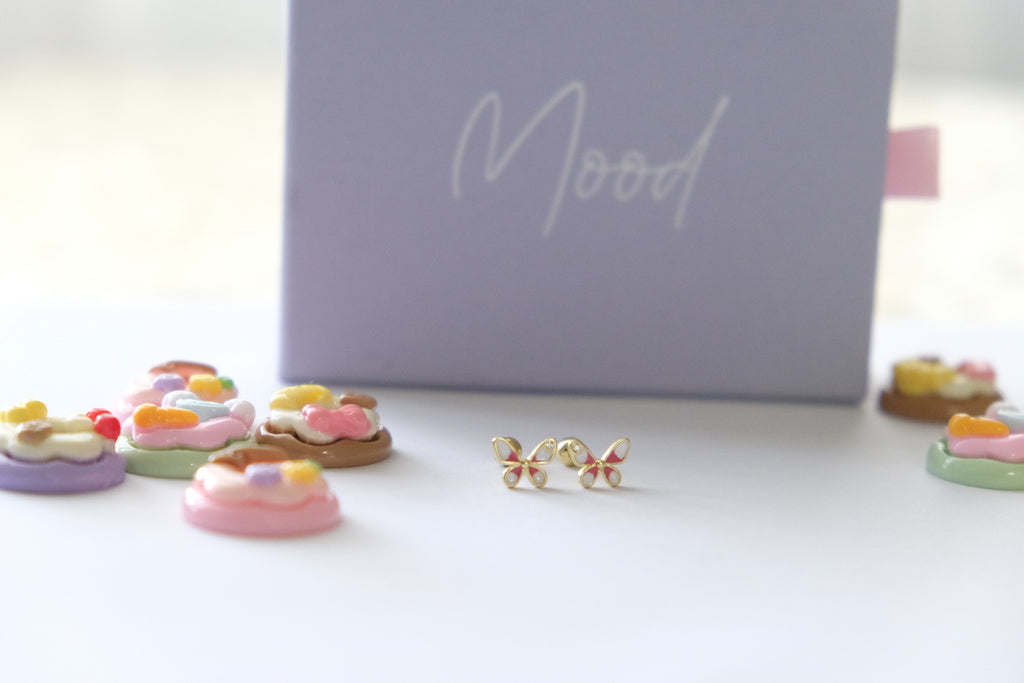 A pair of small gold butterfly stud earrings with pink and yellow enamel details, displayed on a white surface in front of a lavender box labeled 'Mood'.