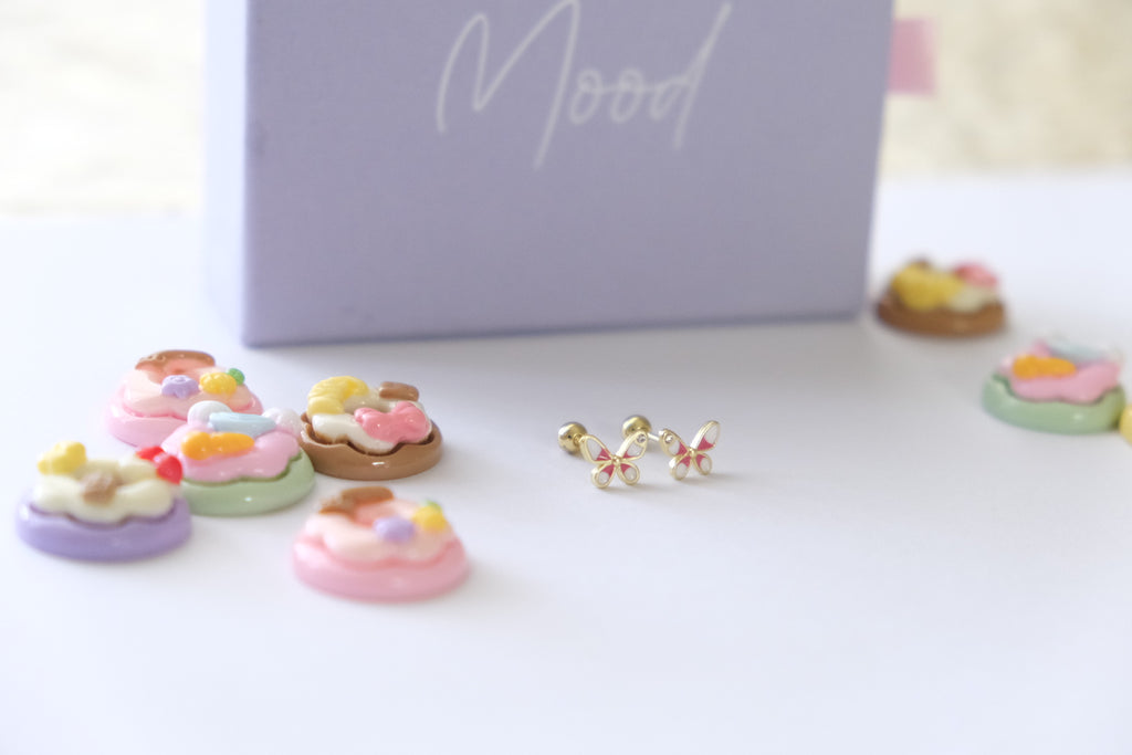 A pair of small gold butterfly stud earrings with pastel pink, yellow, and white details, displayed on a white surface next to a lavender box labeled 'Mood' with miniature pastel cupcake charms.