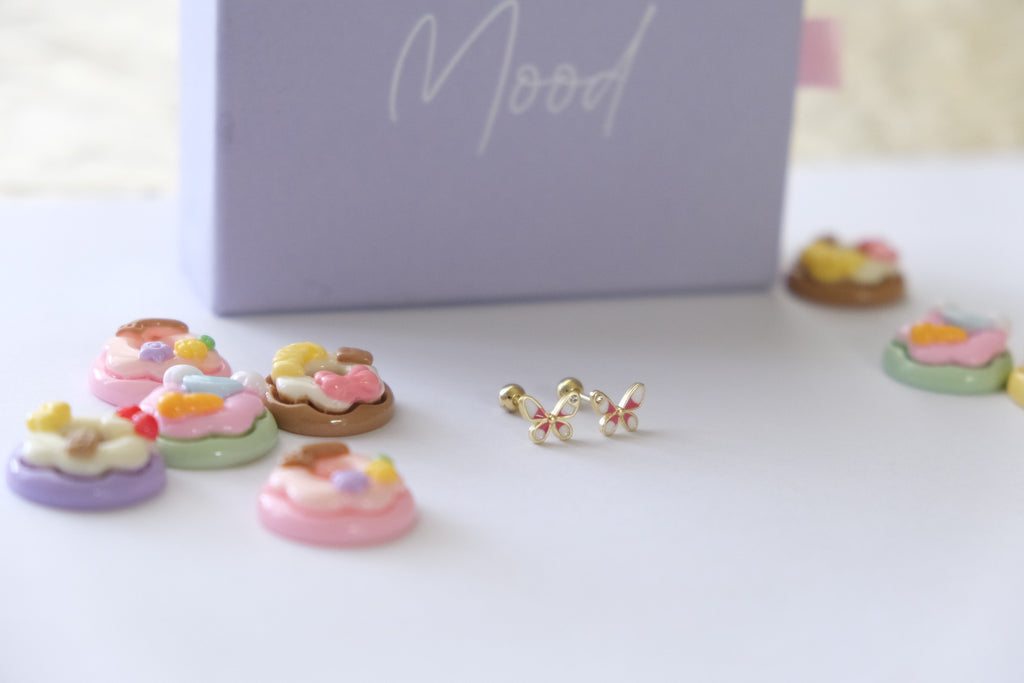 A pair of gold butterfly stud earrings with pastel pink, yellow, and green accents, displayed on a white surface next to a lavender box labeled 'Mood' with miniature cupcake charms.