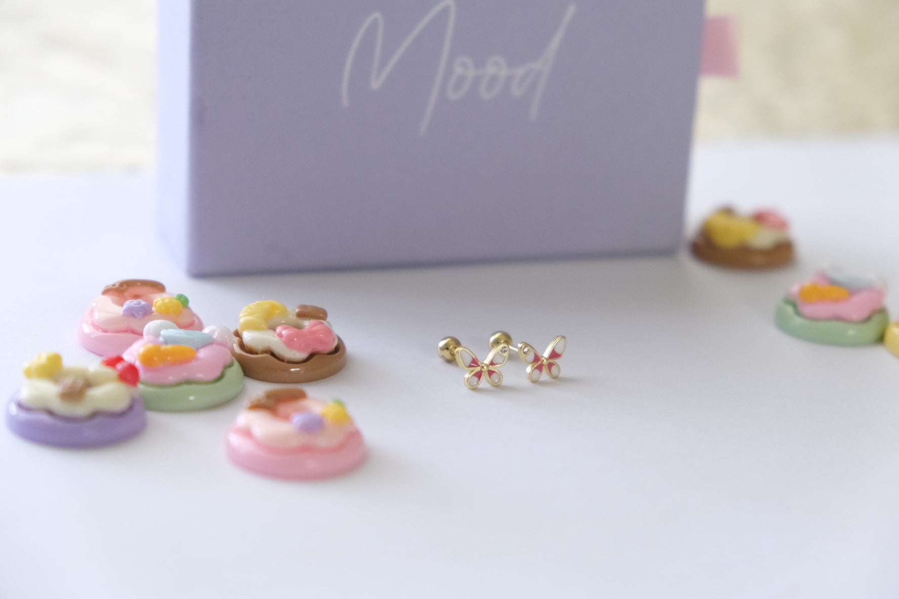 A pair of gold butterfly stud earrings with pastel pink, yellow, and green accents, displayed on a white surface next to a lavender box labeled 'Mood' with miniature cupcake charms.