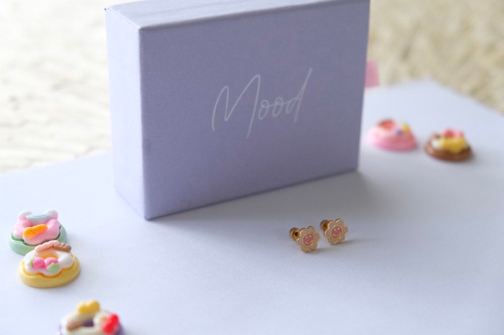 Small decorative earrings on a white surface with a 'Mood' branded box in the background.