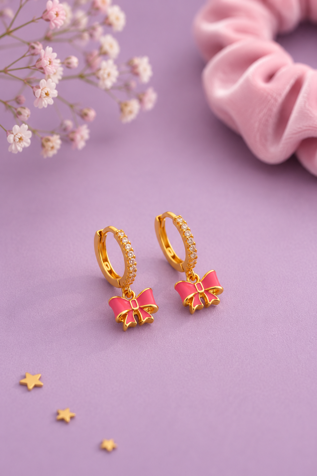 A pair of gold huggie hoop earrings with small clear stones along the top and dangling pink bow charms, displayed on a lavender surface with decorative flowers, a pink scrunchie, and small star accents.