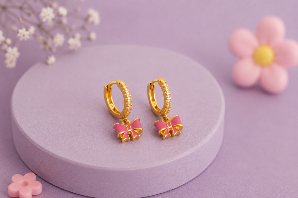 A pair of gold huggie hoop earrings with dangling pink bow charms, displayed on a round lavender pedestal with soft decorative flowers in the background.
