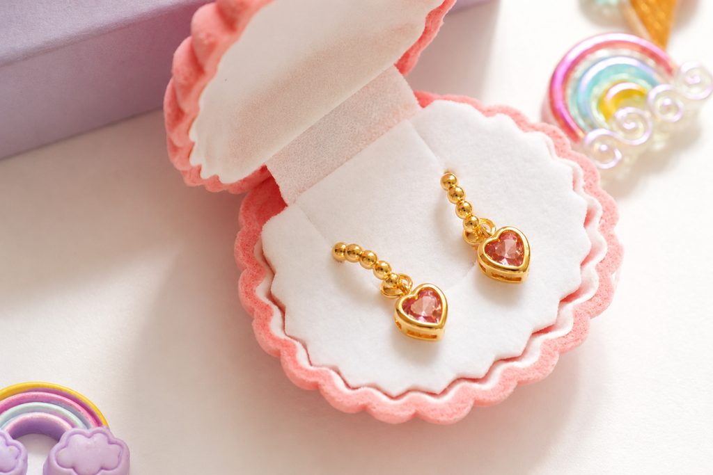 Gold heart-shaped dangle earrings with pink heart charms, displayed in a pink scalloped jewelry box on a white surface.
