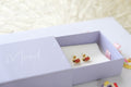 A pair of small gold-tone earrings with red and green ladybug details displayed inside an open lavender gift box with the lid printed with the word 'Mood'.
