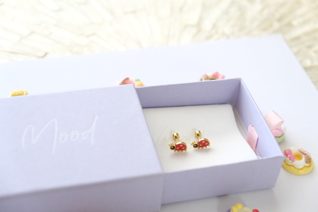 A pair of small gold-toned earrings with red and pink enamel details displayed inside an open lavender jewelry box. The box lid has the word 'Mood' written on it in white script, and the scene is softly lit with a light background.
