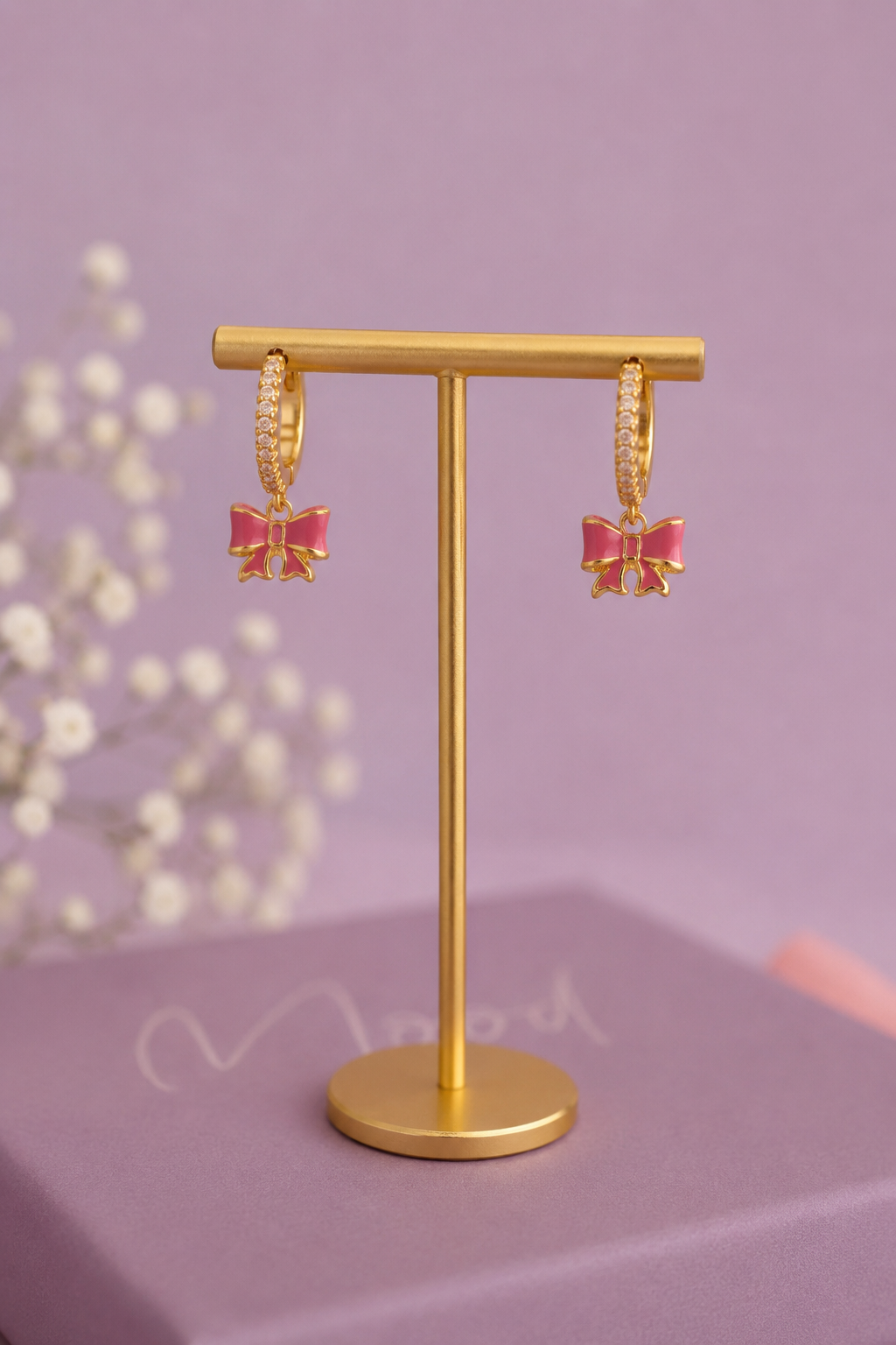 Gold hoop earrings with dangling pink bow charms, displayed on a gold stand against a soft lavender background.
