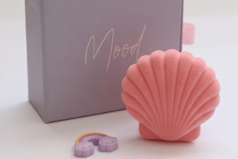 A pink seashell-shaped product displayed next to a lavender-gray box with the word 'Mood' in script, with a small purple rainbow accessory in front on a clean white background.