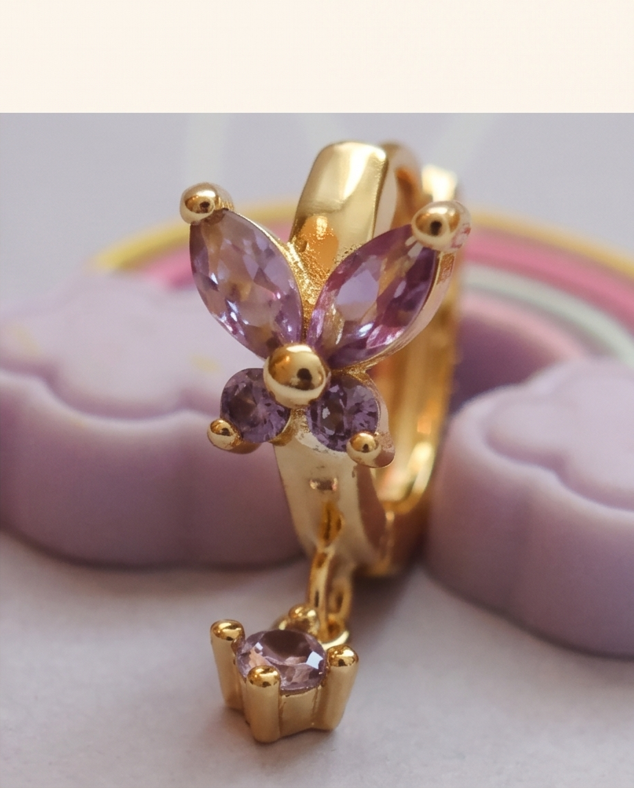 Gold butterfly-shaped earrings with purple gemstones, featuring a dangling crown-shaped charm, displayed on a soft lavender background.