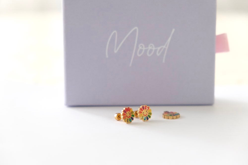 A pair of small gold-toned stud earrings with colorful floral and geometric enamel details, displayed in front of a lavender jewelry box with the word 'Mood' on it.