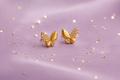 A pair of gold butterfly stud earrings adorned with small clear crystals, displayed on a soft lavender satin fabric with scattered gold glitter.