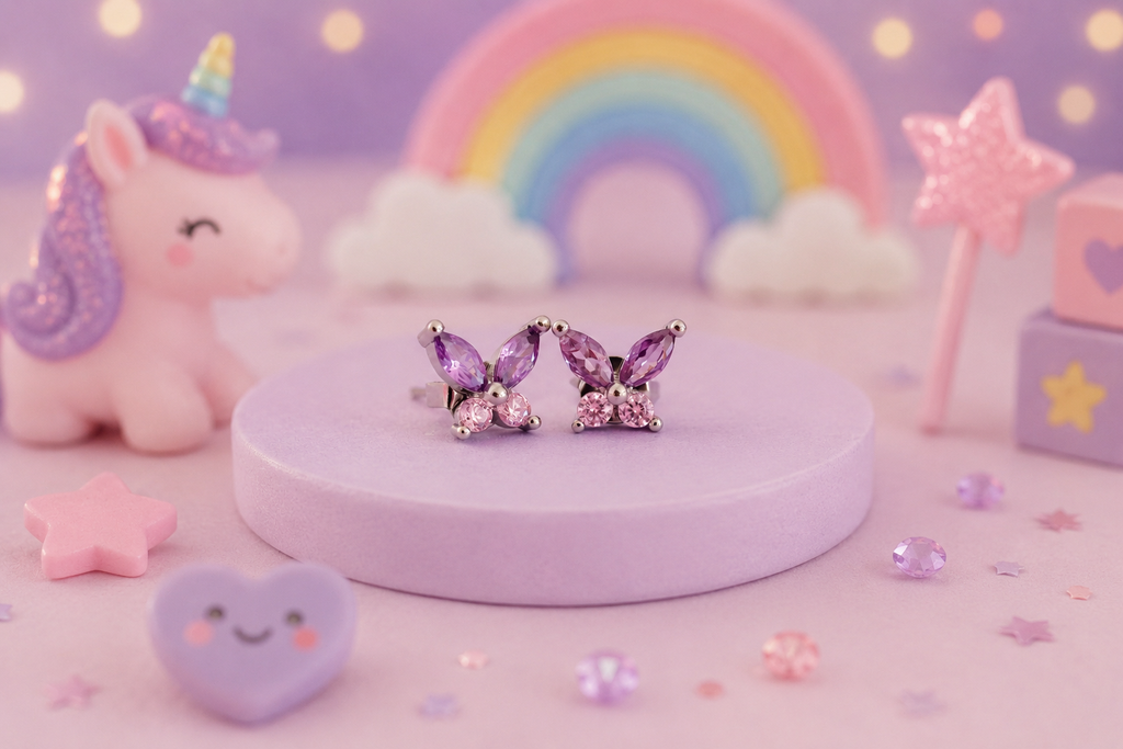 A pair of butterfly-shaped stud earrings with purple marquise-cut rhinestones and silver settings, displayed on a round lavender pedestal. The background is a whimsical, pastel-themed setup featuring a pink unicorn, a rainbow, clouds, and star accessories.