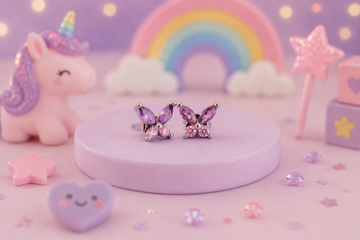 A pair of butterfly-shaped stud earrings with purple marquise-cut rhinestones and silver settings, displayed on a round lavender pedestal. The background is a whimsical, pastel-themed setup featuring a pink unicorn, a rainbow, clouds, and star accessories.