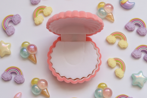 A pastel pink clamshell-shaped compact or case sits open in the center, surrounded by scattered cute charms and accessories including rainbow, moon, star, and ice cream cone shapes in soft pink, purple, yellow, and iridescent tones on a white background.